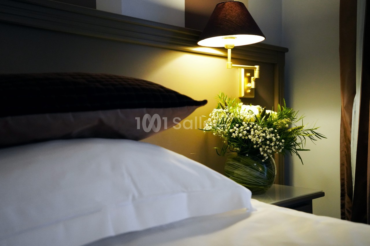 Lit double aux draps blancs, chevet éclairé par une applique dorée et un bouquet de fleurs blanches dans un vase en verre.