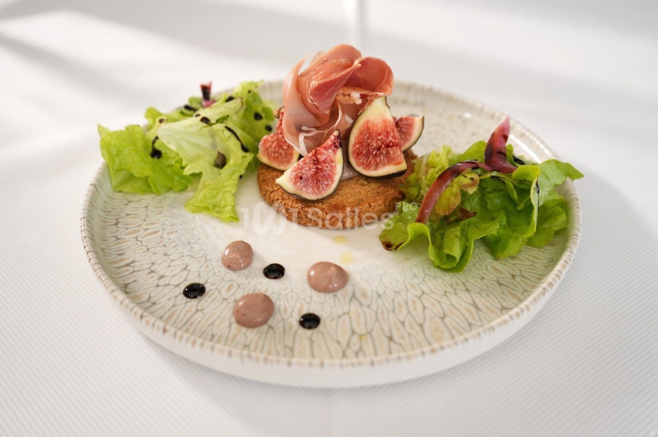 Assiette dressée sur fond blanc : galette croustillante surmontée de tranches de jambon cru et de quartiers de figue, entourée de salade verte, de petites touches de sauce et de filets de glaçage balsamique.