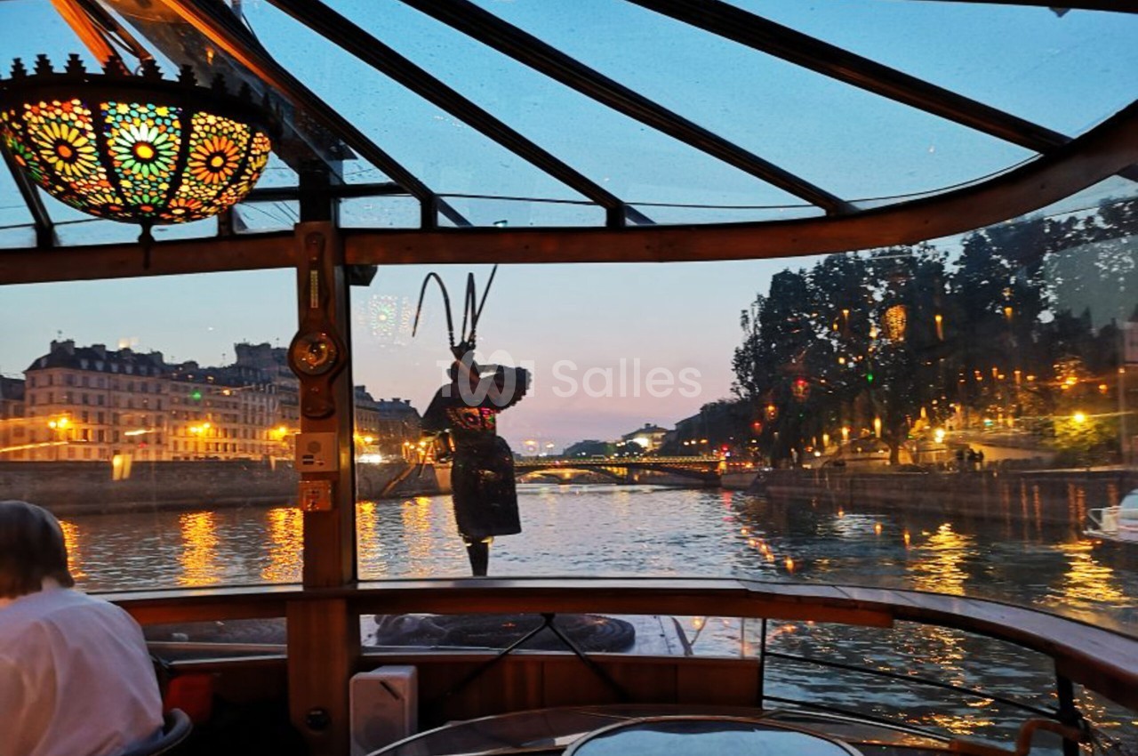 Salle vitrée d'un bateau sur la Seine au crépuscule, avec lustre à vitraux et personnage en costume sombre.