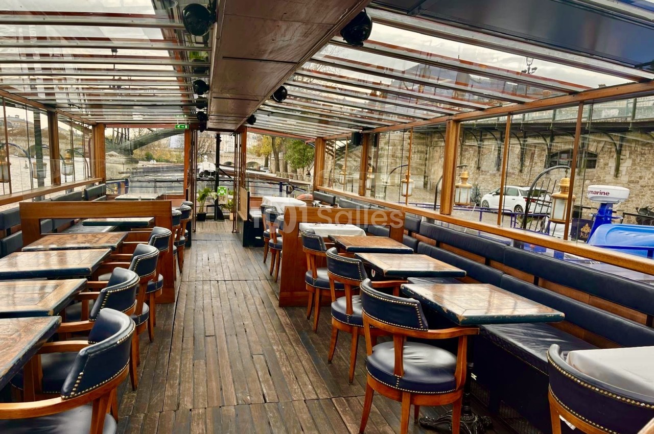 Intérieur vitré d'un bateau : tables au plateau sombre, banquettes et fauteuils en cuir bleu foncé, vue sur le quai.