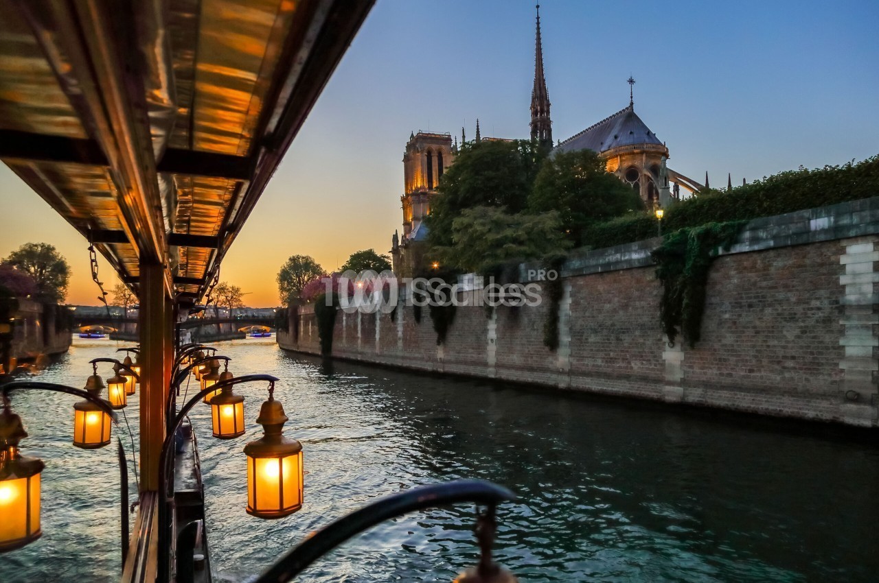 Vue depuis un bateau éclairé de lanternes sur la Seine au crépuscule, avec le chevet d'une cathédrale gothique et un mur de pierre longeant le fleuve. Wait: the instruction says don't start with "Vue de". Let me rewrite. Depuis un bateau aux lanternes