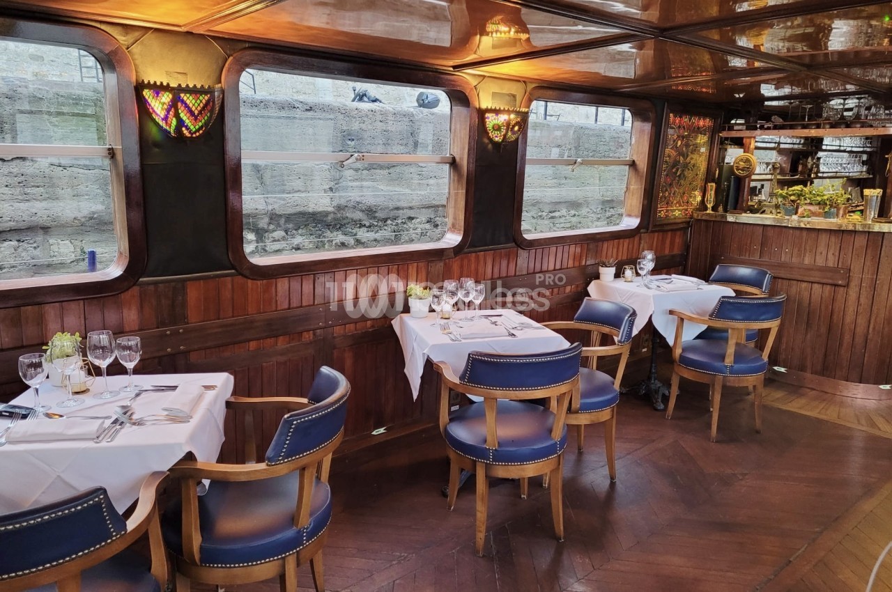Salle de restaurant à bord d'un bateau aux boiseries sombres, avec tables dressées de nappes blanches, fauteuils bleus à clous dorés et grandes fenêtres donnant sur un quai en pierre.