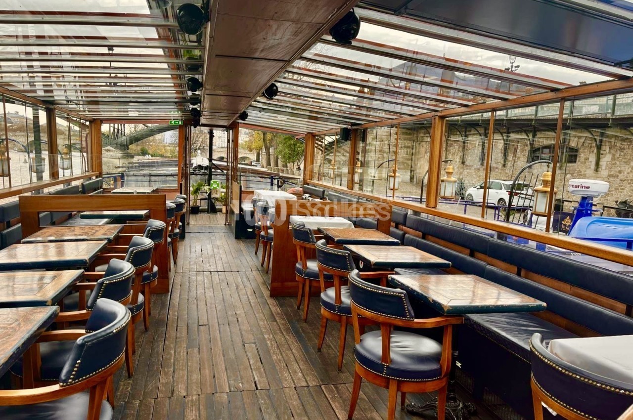 Salle de bateau-restaurant couverte d'un toit vitré, avec tables en bois aux plateaux usés, banquettes et fauteuils en cuir bleu marine, vue sur les quais.