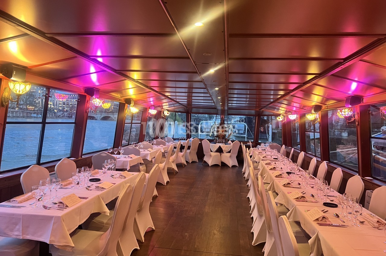 Salle de bateau-restaurant aux longues tables dressées de nappes blanches et chaises habillées de housses blanches, éclairée de lumières roses et ambrées.