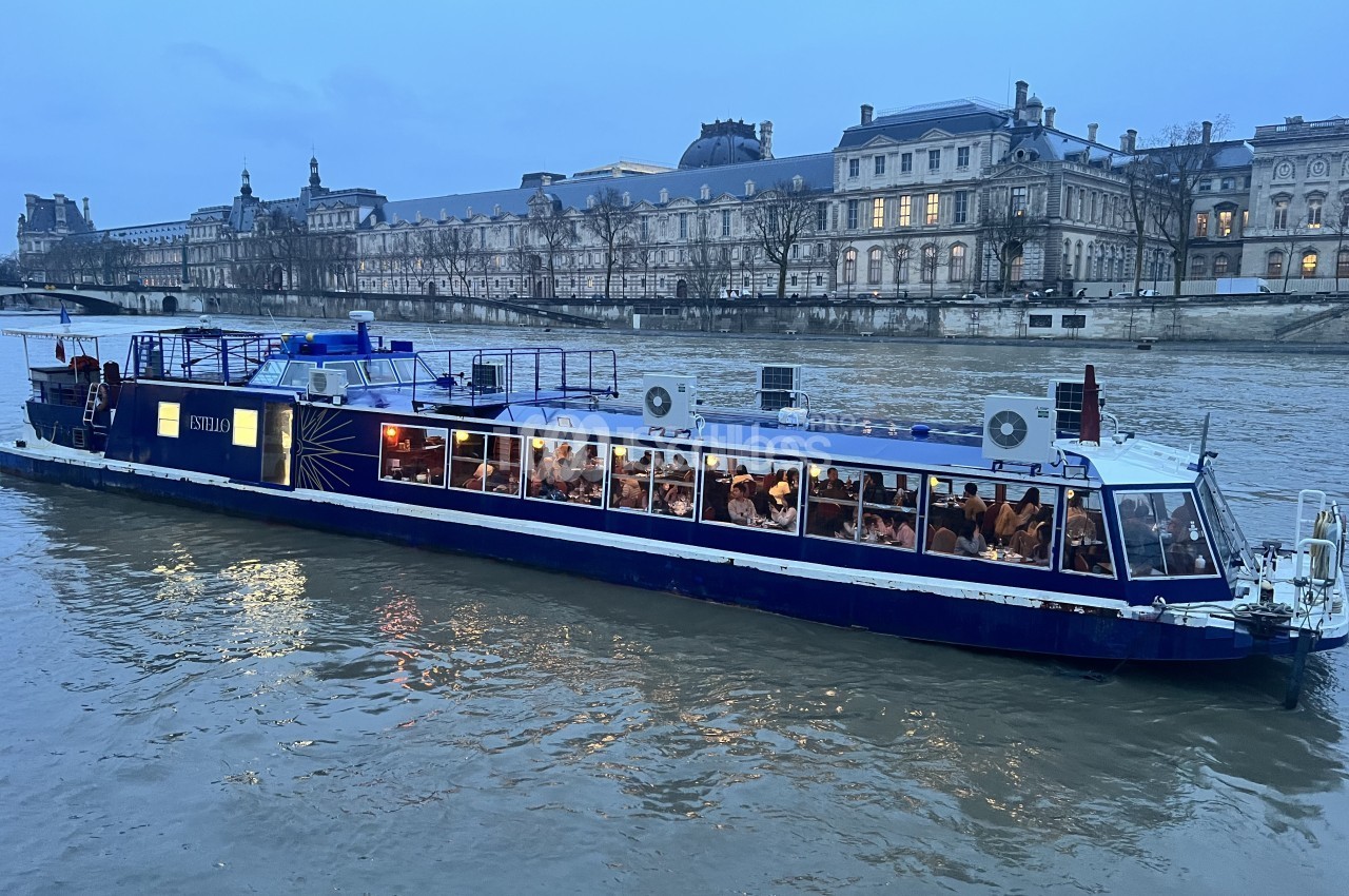 Bateau-restaurant bleu sur la Seine au crépuscule, avec des convives attablés derrière de grandes baies vitrées et des bâtiments historiques en arrière-plan.
