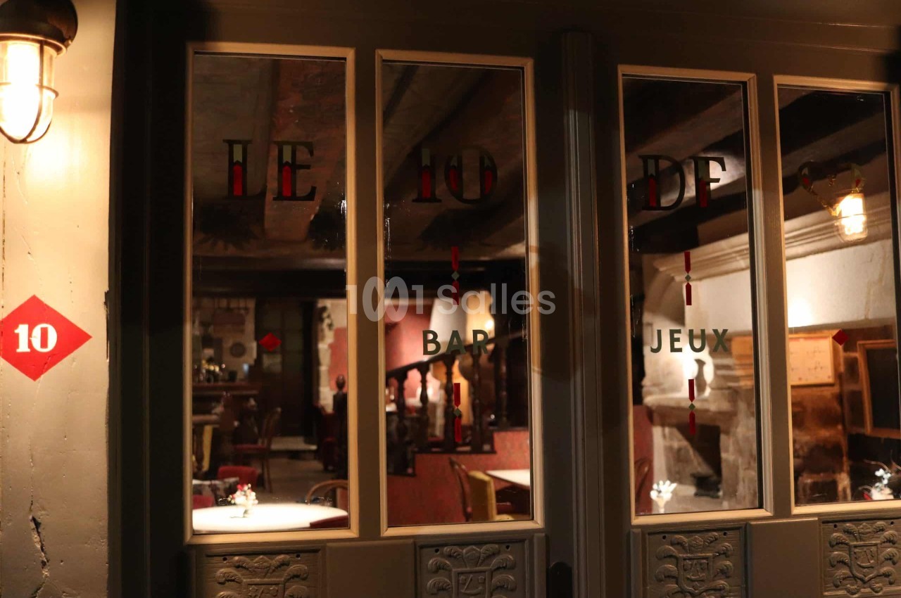 Façade à grandes vitres encadrées de gris foncé, le soir, donnant sur une salle aux tables dressées et à l'éclairage chaud.