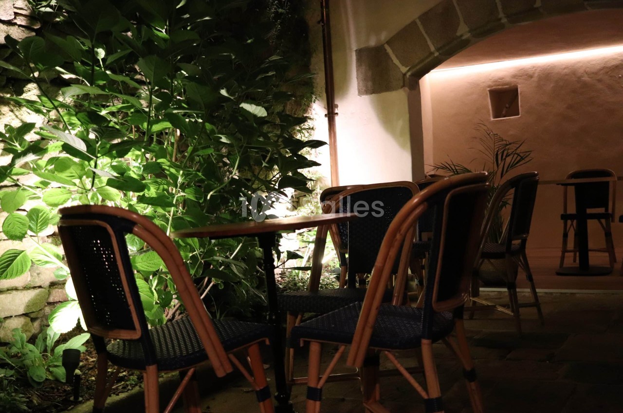 Terrasse nocturne en pierre sous une voûte éclairée, avec tables rondes et chaises en rotin bleu marine, bordée d'une haie de feuillage vert.