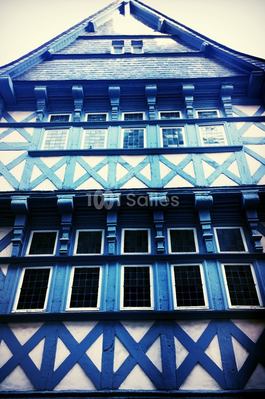 Façade à colombages peinte en bleu, vue en contre-plongée, avec plusieurs rangées de fenêtres à petits carreaux et un toit en ardoise.