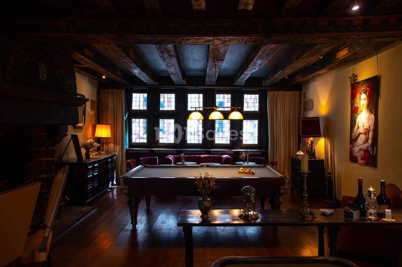 Salon aux poutres apparentes avec une table de billard au centre, canapés bordeaux sous une rangée de hautes fenêtres à petits carreaux.