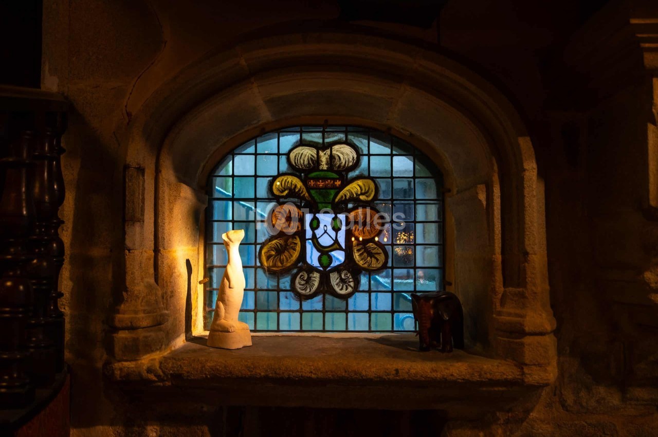 Fenêtre cintrée en pierre à vitrail bleu et jaune représentant un écusson, avec une statuette blanche et une figurine en bois posées sur un rebord.