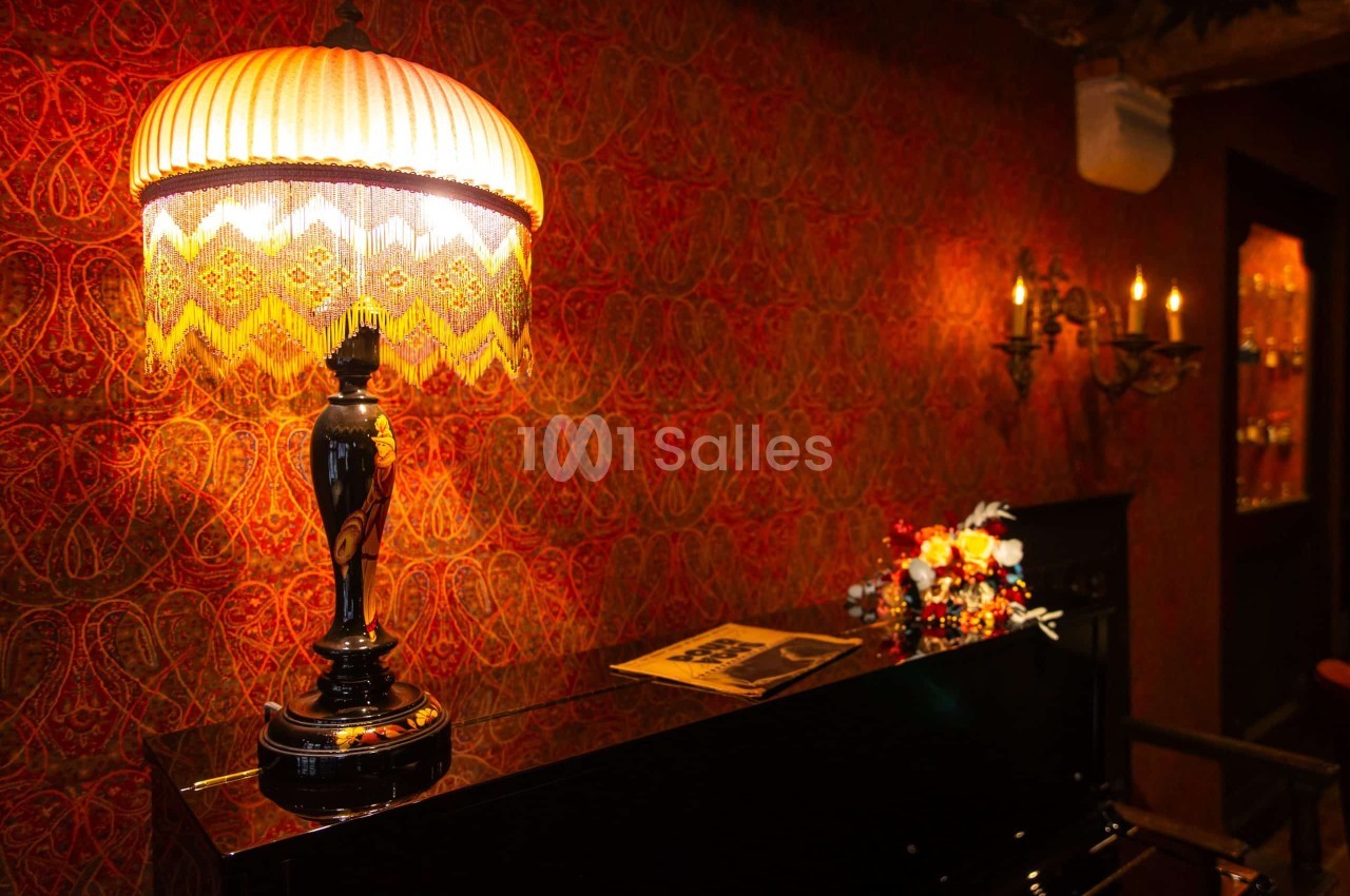 Lampe de table à abat-jour plissé orné de franges dorées, posée sur un meuble laqué noir, devant un mur tapissé de motifs cachemire rouges et chauds.