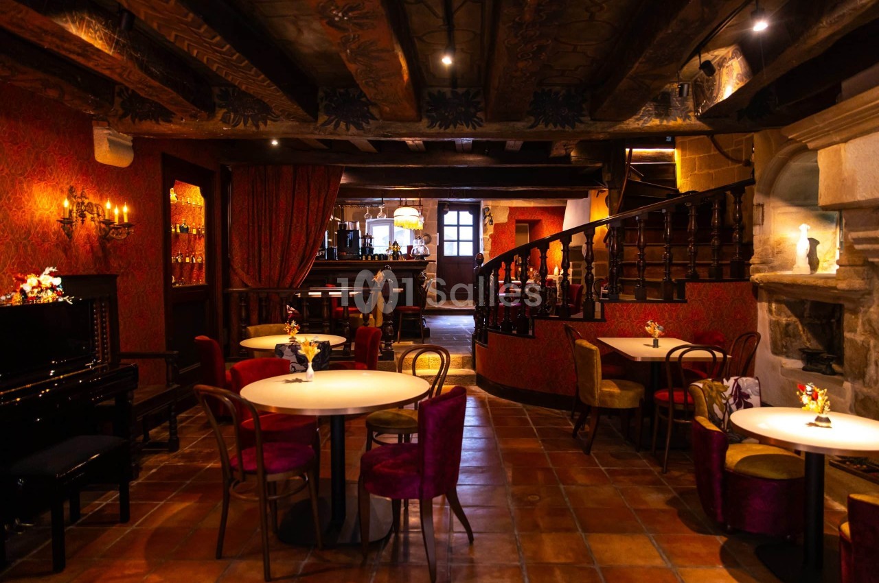 Salle de restaurant aux murs rouges et plafond en poutres de bois sombres, avec tables rondes, chaises au velours bordeaux, un piano à queue et un escalier en bois.