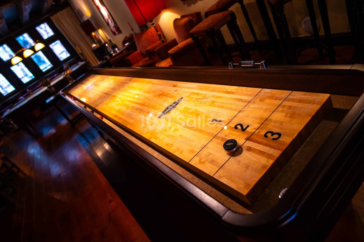 Table de shuffleboard en bois clair aux zones de score numérotées 1, 2 et 3, dans un bar à l'éclairage tamisé avec banquettes en arrière-plan.