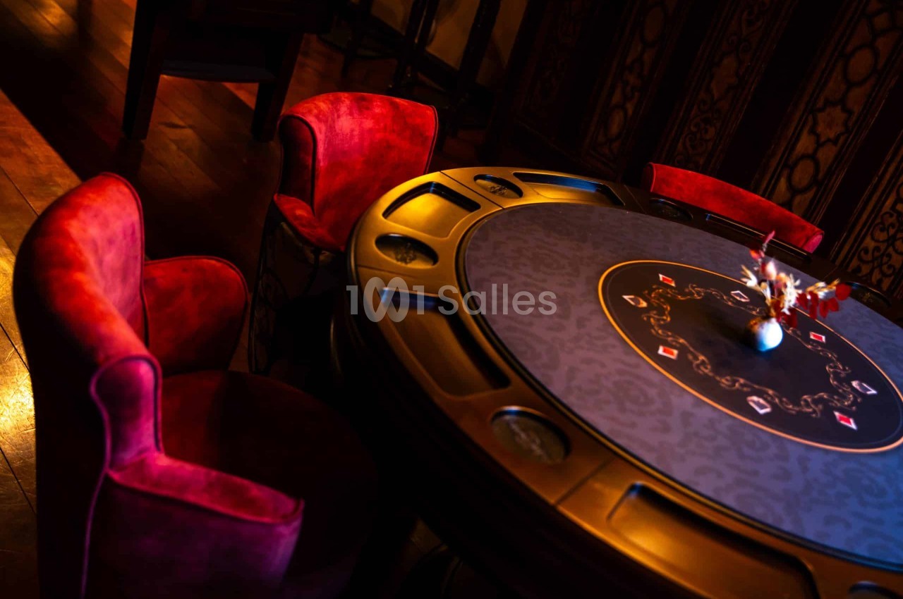 Table ronde au tapis bleu à motifs de cartes, entourée de fauteuils en velours rouge, dans une salle à l'éclairage tamisé.