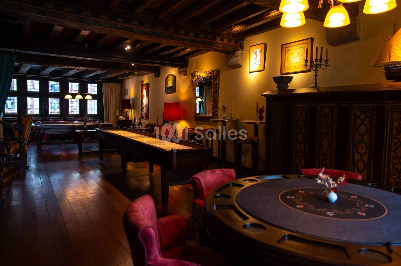 Salle aux poutres apparentes avec table ronde à motifs de cartes, fauteuils en velours rouge et jeu de palets en bois.