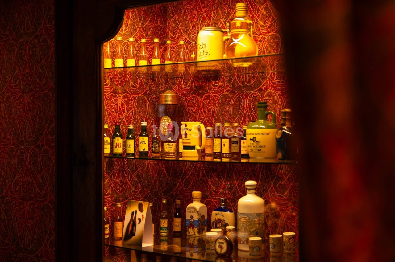 Vitrine éclairée en ambre présentant une collection de bouteilles de whisky miniatures, carafes en grès et chopes sur plusieurs étagères en verre.