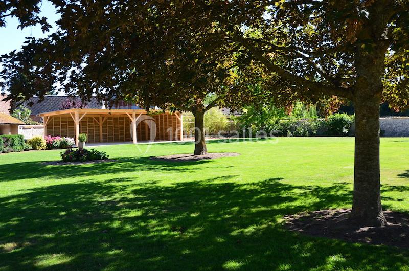 Pelouse verte avec arbres ombragés et abri en bois au fond, entourée de haies et d'un mur en pierre.