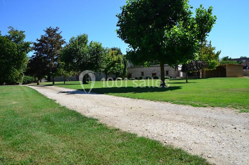 Allée de gravier traversant une pelouse verdoyante avec des arbres et des bâtiments en pierre à l'arrière-plan.