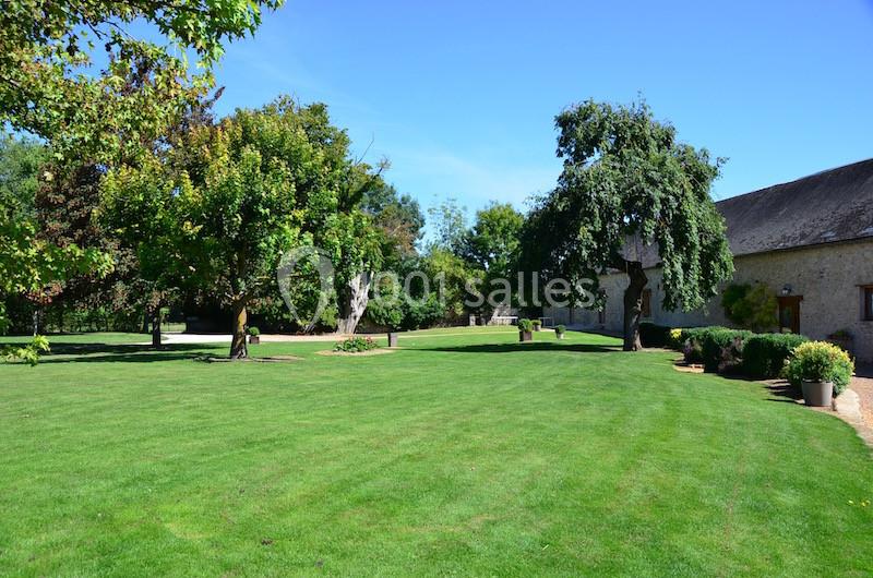 Jardin verdoyant avec pelouse, arbres et bâtiment en pierre sous un ciel bleu clair.