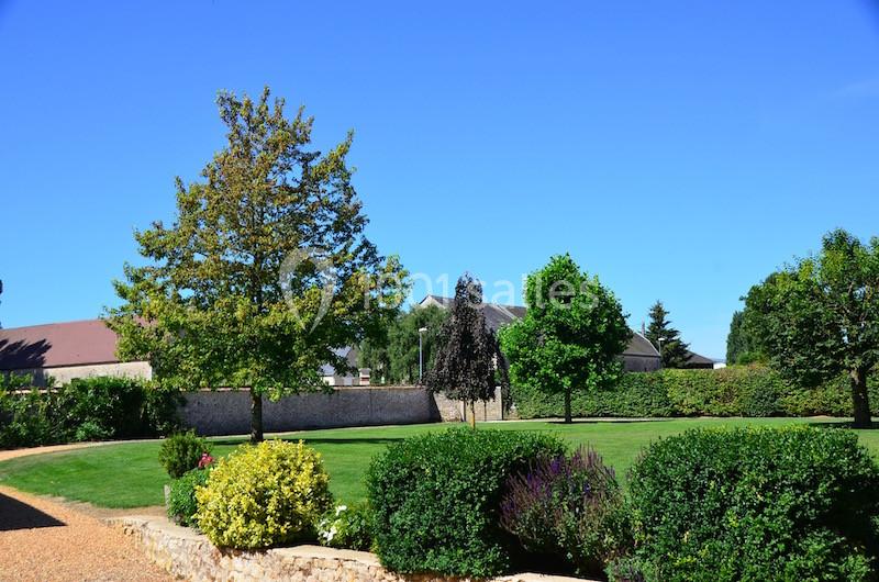 Jardin verdoyant avec pelouse, arbustes et arbres sous un ciel bleu, entouré de bâtiments et d'un mur en pierre.