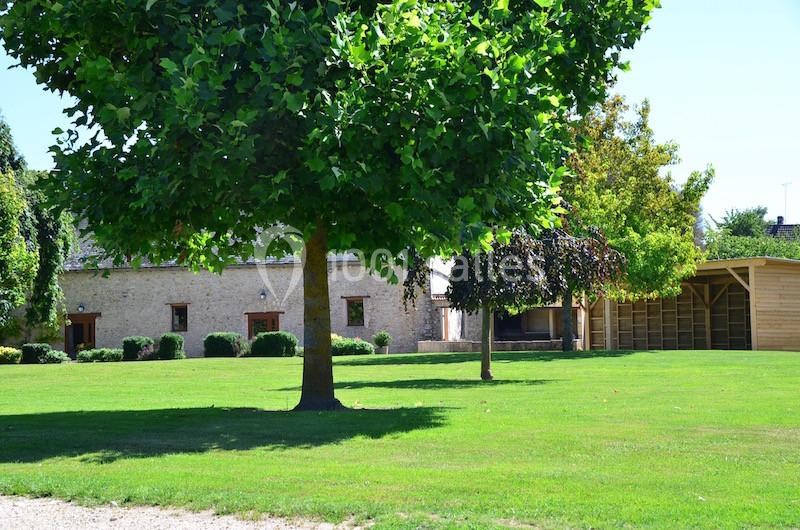 Pelouse verdoyante avec un grand arbre au premier plan, bâtiment en pierre et abri en bois en arrière-plan.