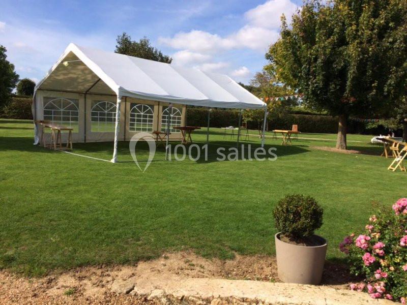 Tente blanche montée sur une pelouse, entourée d'arbres et de tables en bois, sous un ciel dégagé.