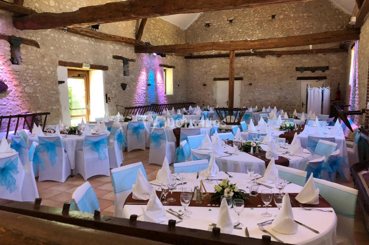 Salle de réception décorée pour un événement, avec tables rondes dressées, nappes blanches et nœuds bleus sur les chaises.