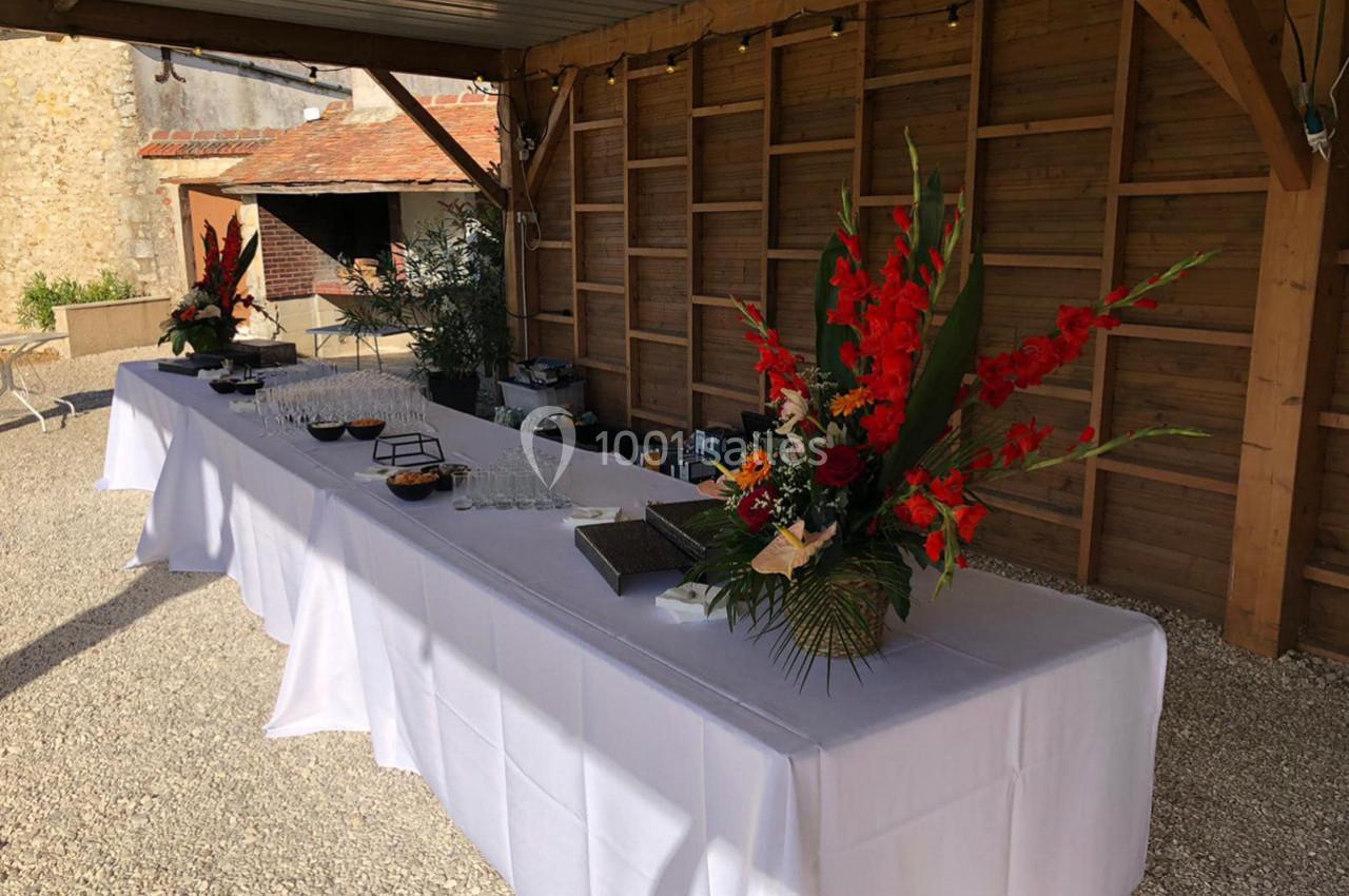 Table dressée sous un abri en bois, décorée de bouquets de fleurs rouges et de verres alignés pour un événement.