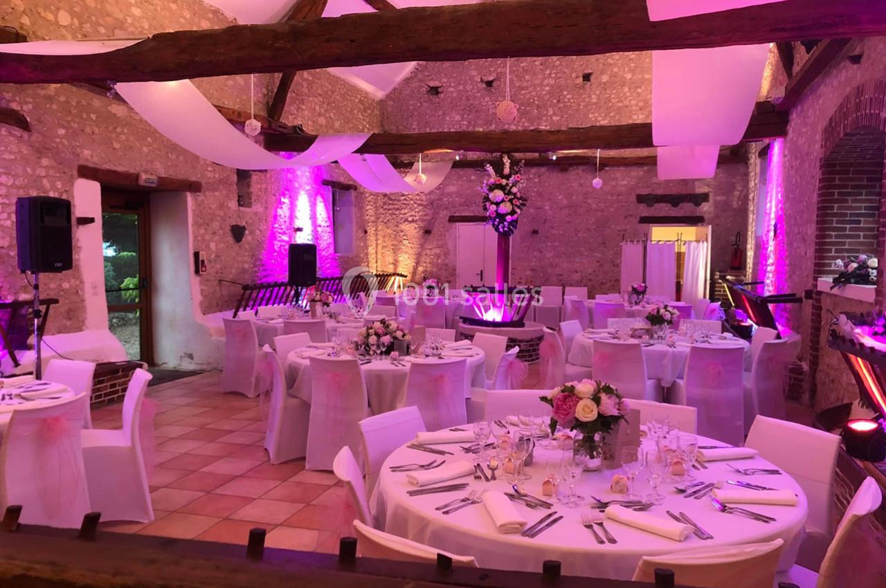 Salle de réception décorée pour un mariage, avec tables rondes dressées, nappes blanches et éclairage rose.
