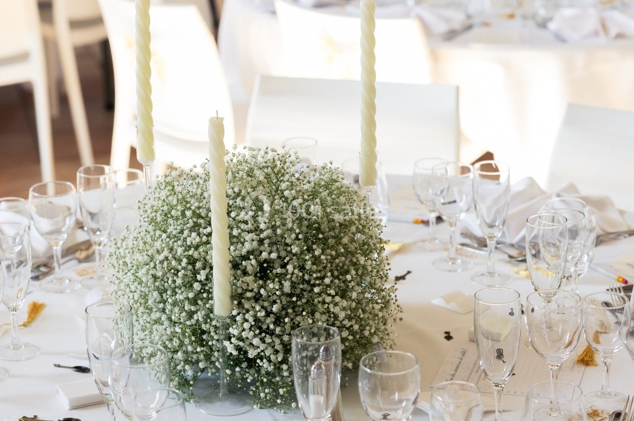 Tables de réception décorées de nappes blanches, bougies et bouquets de gypsophile, avec vaisselle et couverts disposés.