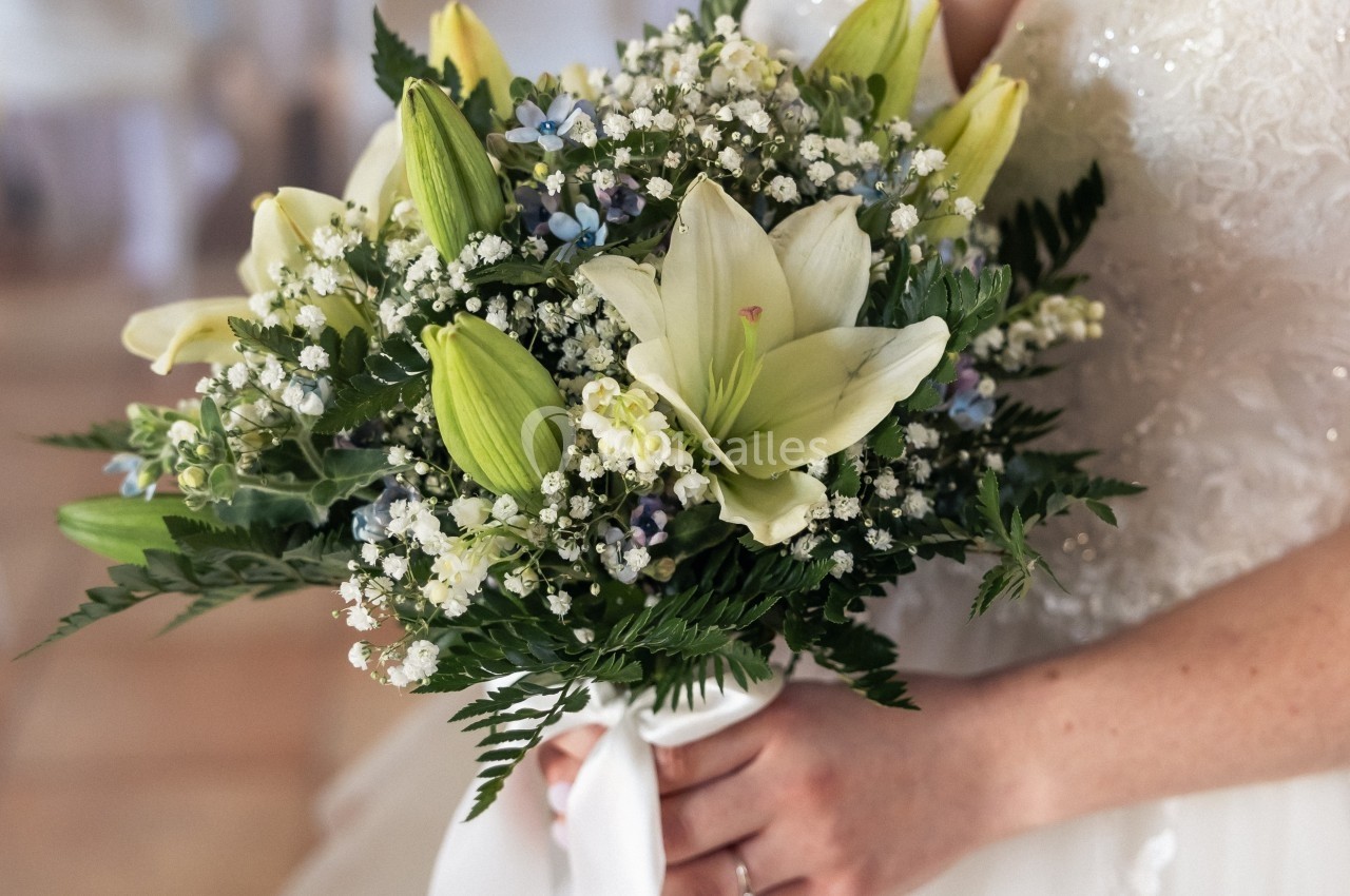 Femme en robe de mariée tenant un bouquet de lys blancs, gypsophiles et feuillage, noué d'un ruban blanc.