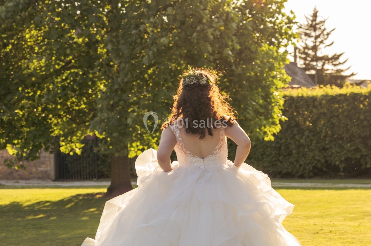 Une femme en robe de mariée volumineuse marche sur une pelouse ensoleillée, entourée d'arbres et de verdure.