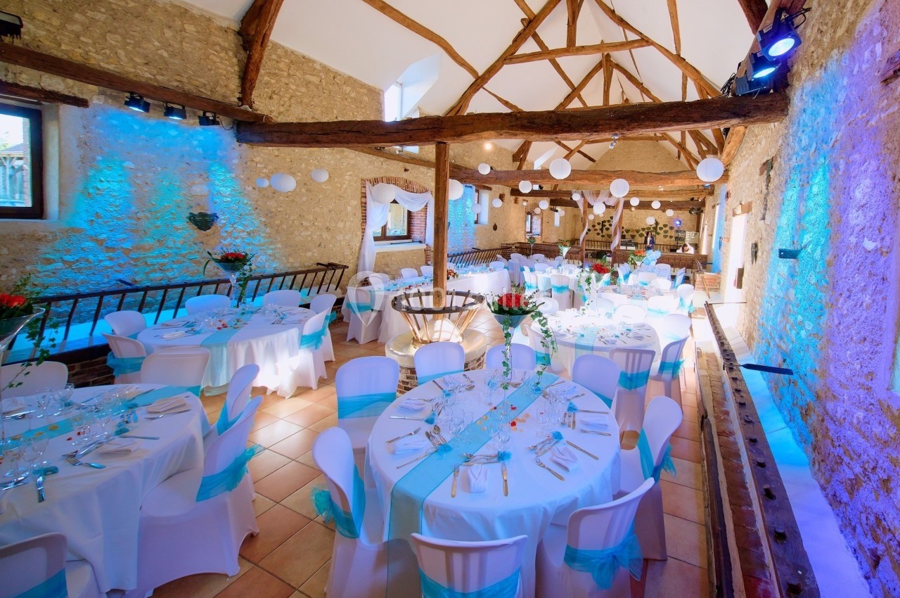 Salle de réception en pierre avec poutres apparentes, tables rondes décorées de nappes blanches et rubans bleus.