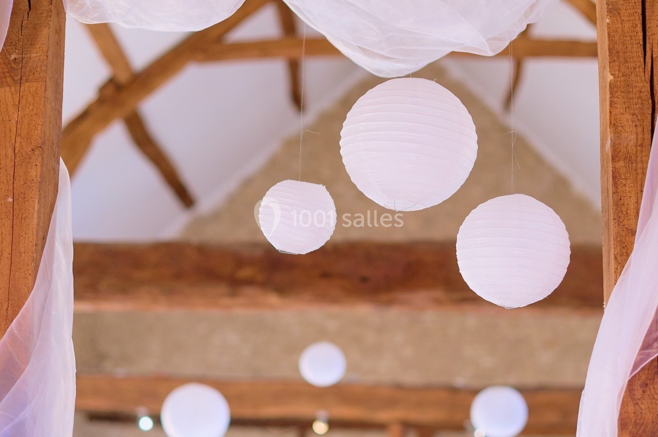 Lampions blancs suspendus sous une charpente en bois, décorés de voilages translucides dans un espace lumineux.