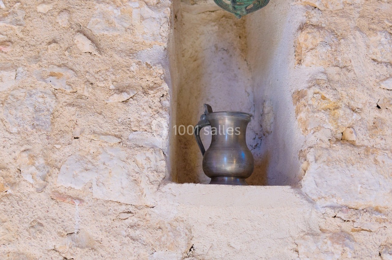 Une cruche en métal posée dans une niche murale en pierre, encadrée par deux plaques anciennes en métal.