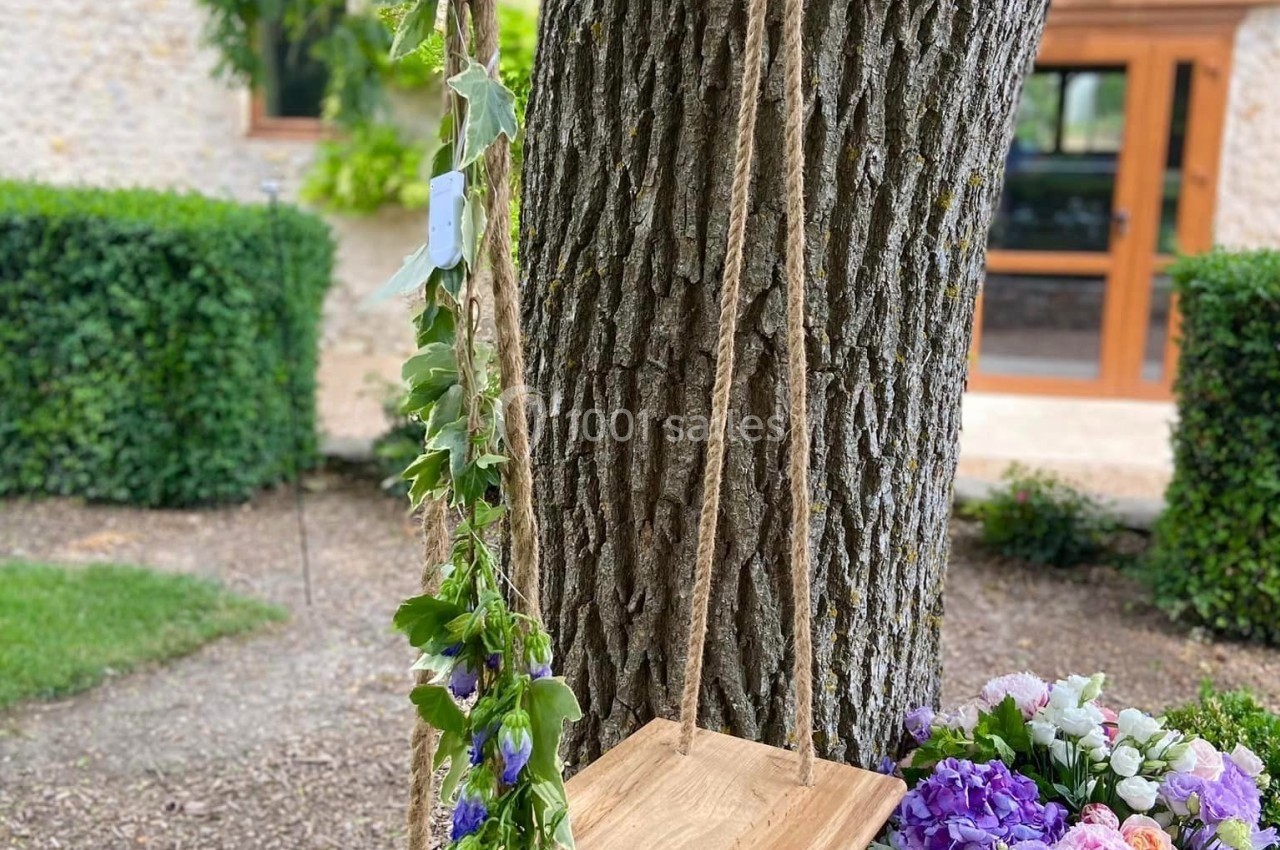 Balançoire en bois suspendue à un arbre, décorée de fleurs, avec un bouquet coloré posé à côté.