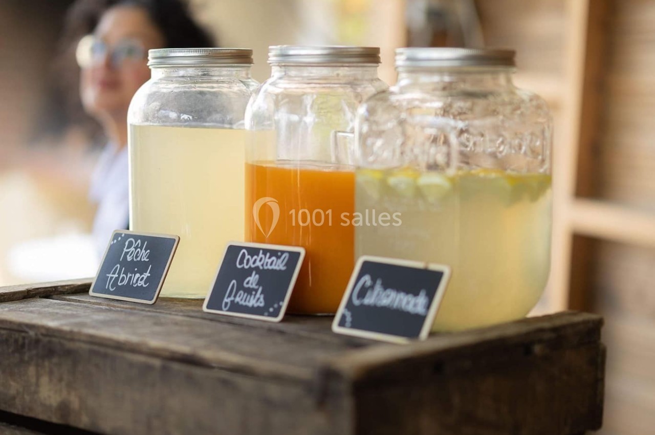 Trois jarres en verre contenant des boissons variées posées sur une caisse en bois, avec des étiquettes écrites à la main.