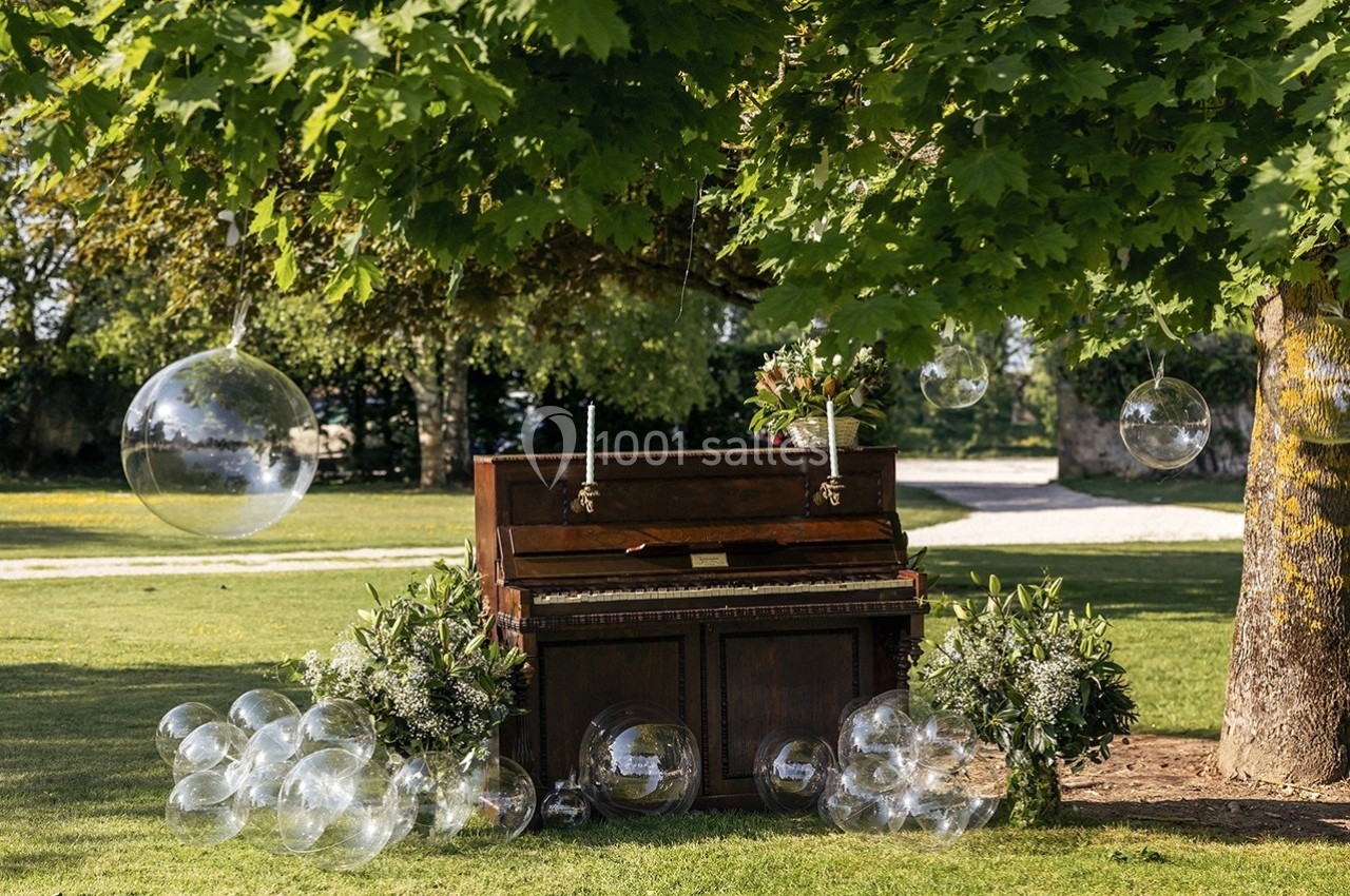 Piano ancien décoré de fleurs et entouré de bulles transparentes, installé sous un arbre dans un jardin.