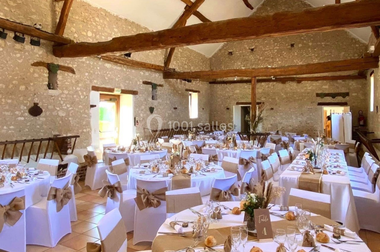 Salle de réception en pierre avec poutres apparentes, tables décorées pour un événement, nappes blanches et chaises ornées…
