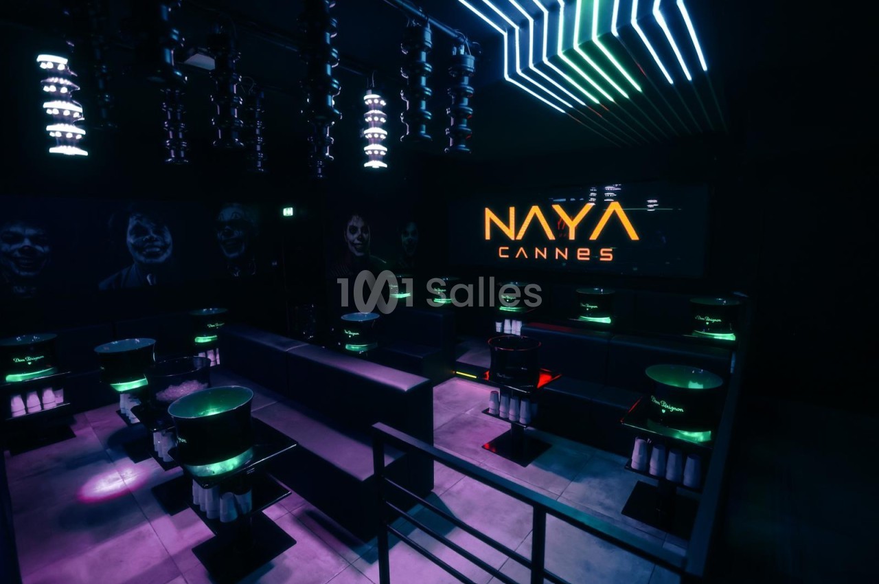 Salle de club en pénombre avec banquettes sombres, tables lumineuses vertes et violettes, éclairage néon bleu et vert au plafond.