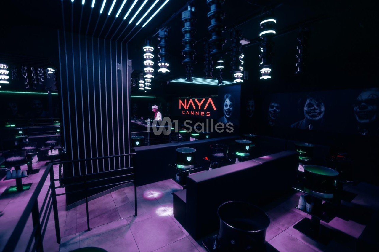 Salle de club aux éclairages violets et verts, avec tables hautes rondes, banquettes sombres et luminaires suspendus, côté bar en fond.