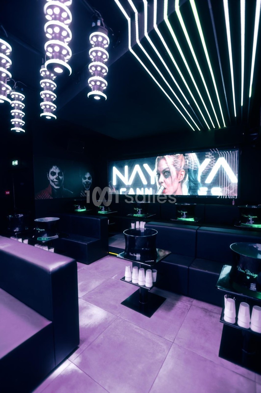 Intérieur de club sombre aux banquettes noires et tables hautes éclairées par des néons verts, avec un grand écran lumineux au mur et des luminaires LED suspendus.