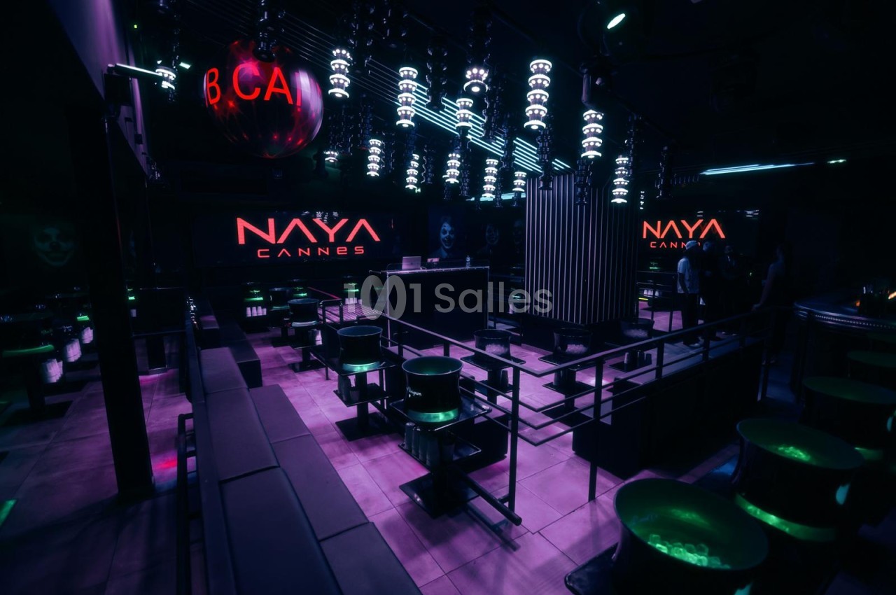 Intérieur de club nocturne éclairé en violet, avec tables hautes rondes à éclairage vert et luminaires au plafond.
