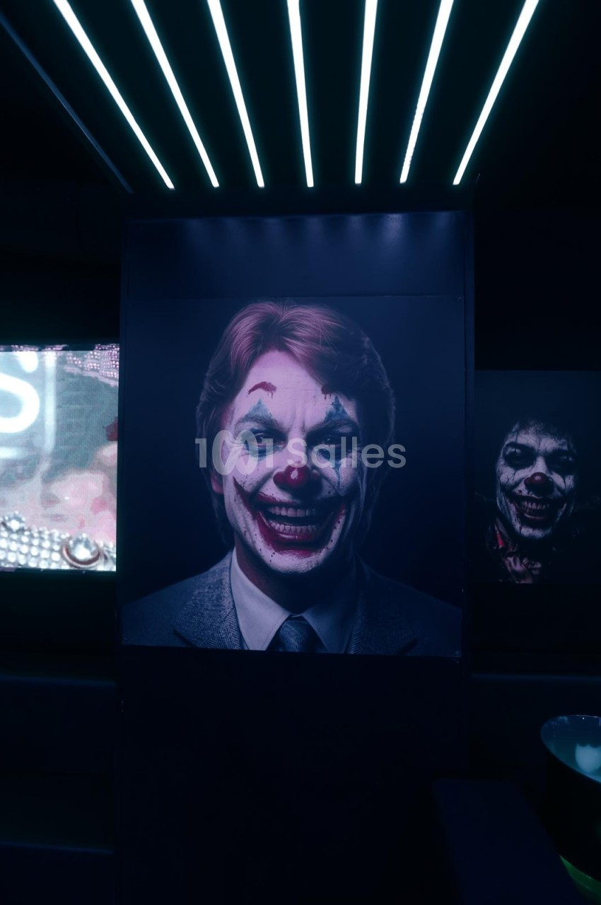 Salle sombre éclairée par des néons au plafond, grand écran affichant un portrait d'homme en costume maquillé en clown.
