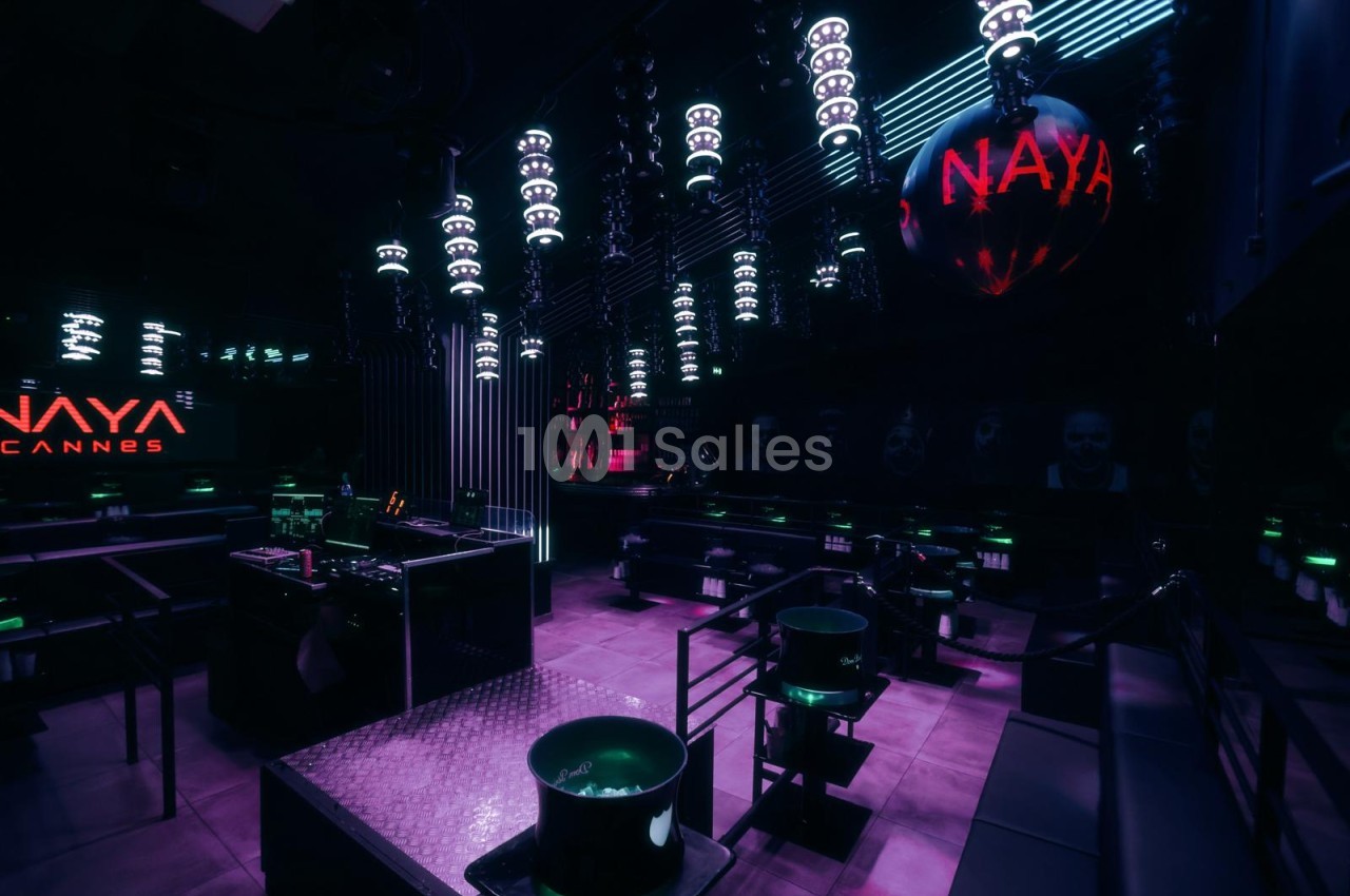 Club à l'ambiance sombre éclairée de violet, avec lampes suspendues, banquettes et seaux à glace sur tables basses.