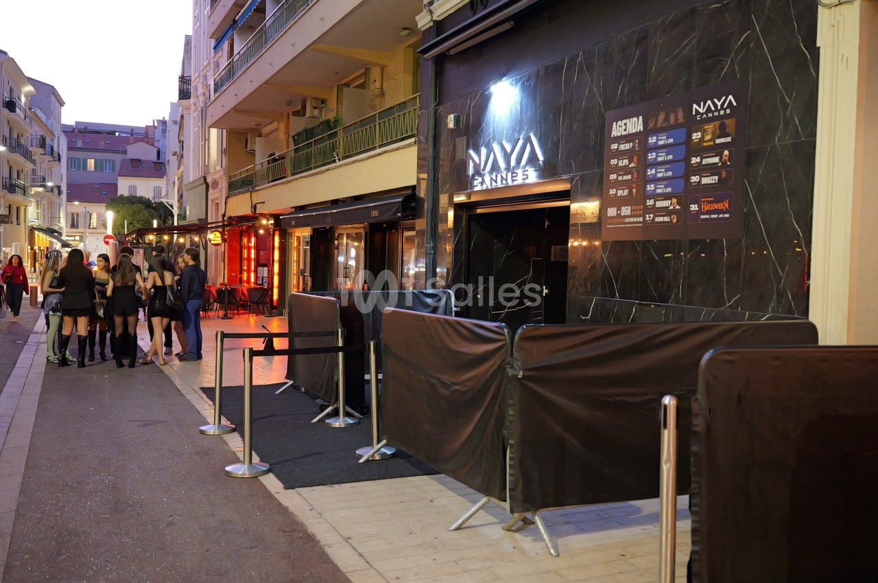Façade d'un club de nuit en soirée, avec entrée en marbre noir, barrières métalliques et files de personnes sur le trottoir. Let me check constraints: no "Photo de", one sentence, under 125 chars. Count: roughly 125? "Façade d'un club de nuit en soirée