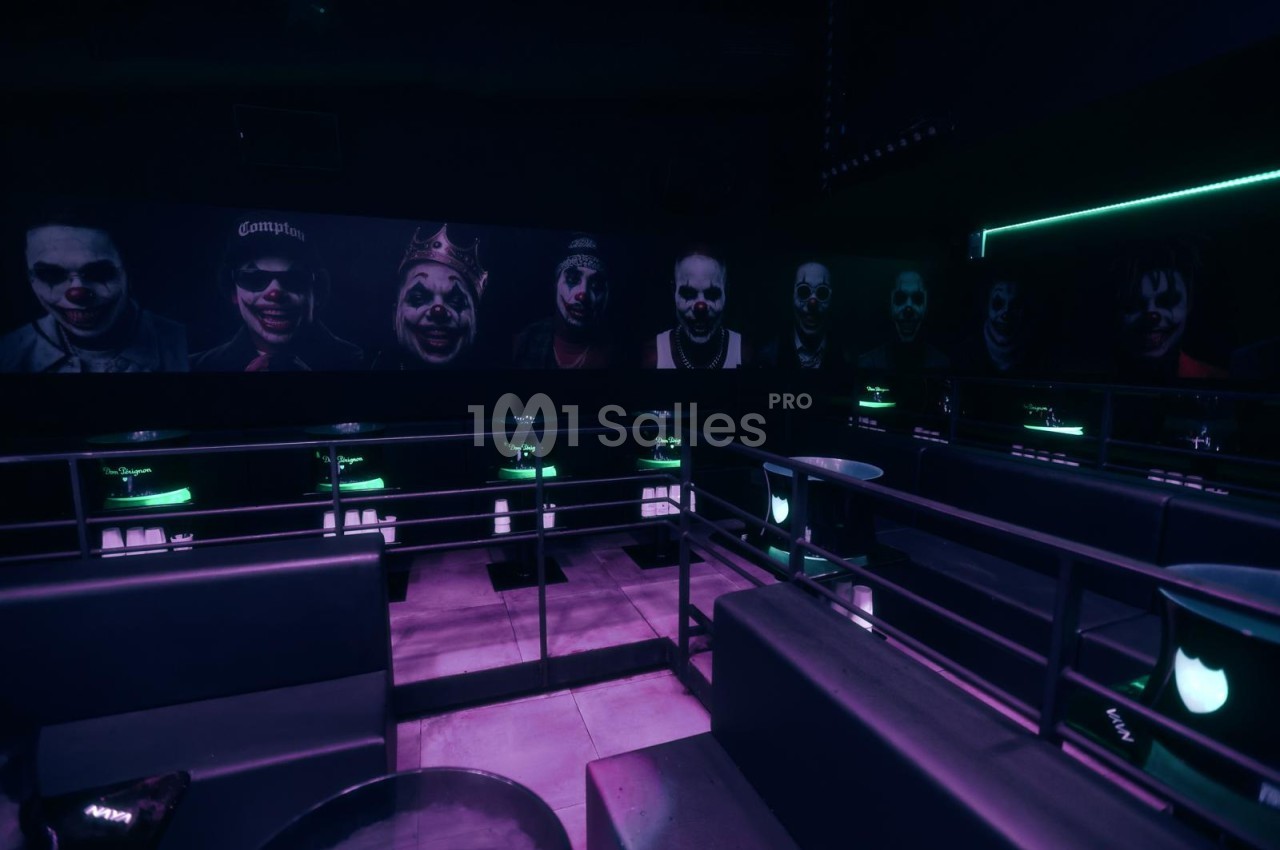 Salle de club en éclairage violet et vert, banquettes sombres, tables rondes et grand mur de portraits masqués.