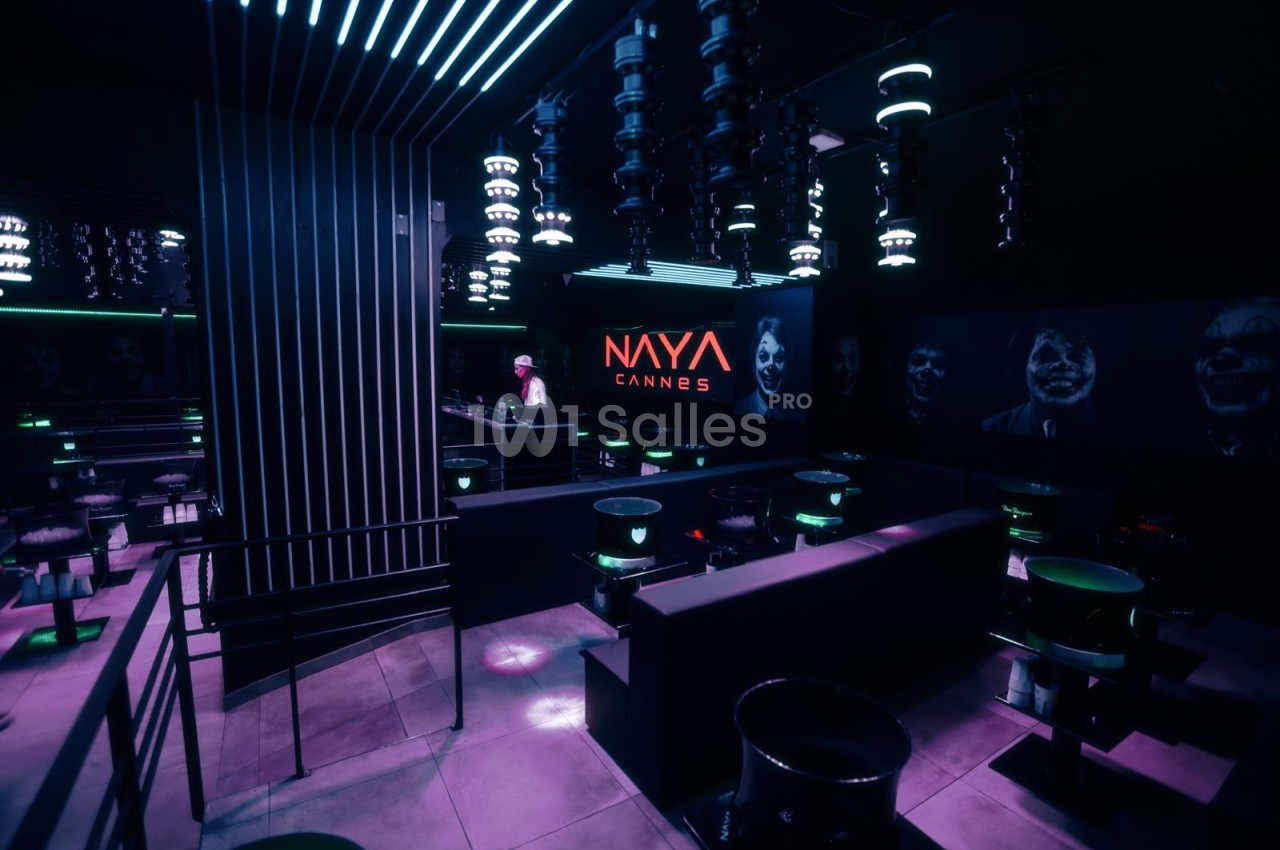 Salle de club aux éclairages violets et verts, avec tables hautes rondes, banquettes sombres et luminaires suspendus, côté bar en fond.