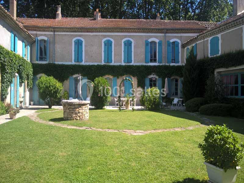 Cour intérieure d'une maison en pierre avec volets bleus, pelouse, puits central et végétation environnante.