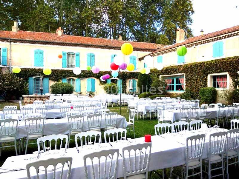 Tables dressées avec nappes blanches et chaises alignées dans une cour extérieure décorée de ballons colorés.